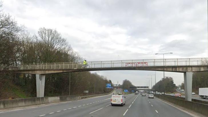 Three lanes of a motorway with a slip road to the left and a blue sign in the distance. There is a white van travelling ahead and cars in the distance too.