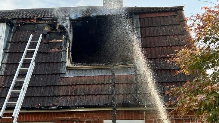 The roof of a house where the force of a fire has blown out a dormer window. A ladder is propped against the roof and a jet of water from a fire hose is being sprayed through the window void and into the building.