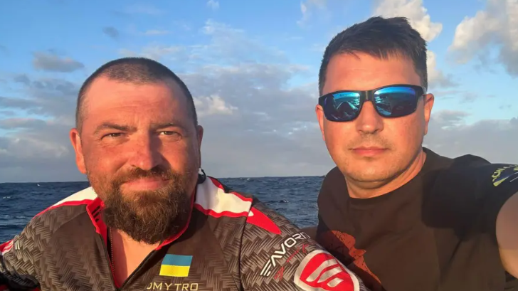 Dmytro Rezvoy, in blue sunglasses and a black T-shirt, and Ivan Havrylko, wearing a shirt with Ukraine's flag on it, in their rowing boat at sea. 