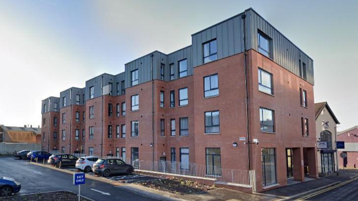 A Google Maps Streetview screengrab of an apartment block on Wardsborough Road, Lisburn on a bright day in 2021.  The four-storey building is red brick with grey panelling or cladding on the top floor.  Several cars are parked outside the flats. 