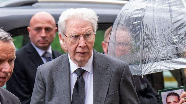 Jim Craih has white hair and wears glasses and a dark grey suit. People around him wear dark clothes and someone is holding a clear umbrella