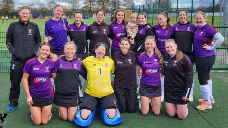 A hockey team comprised of 14 women - mostly wearing club colours of black and purple - assembled in two rows. Half are standing at the back row, one holding a young girl. The others are kneeling in front. To the left, a man stands wearing black sports clothes. The players include a goalkeeper wearing a yellow top and blue knee guards. Everyone is smiling at the camera.