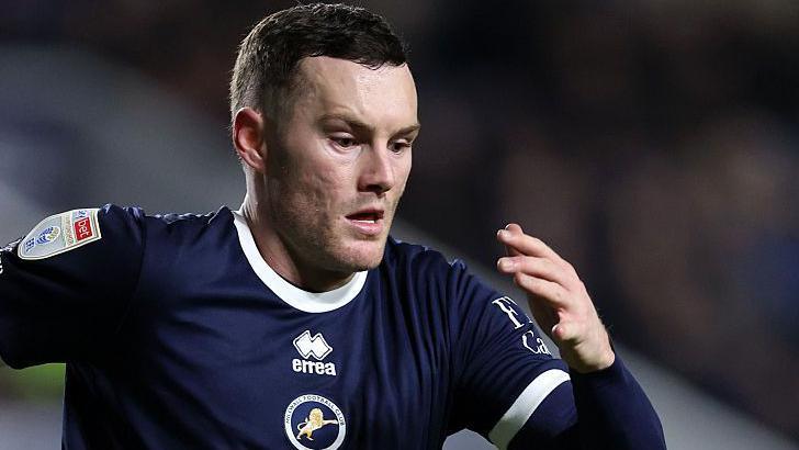 Salford City striker Macaulay Langstaff pictured in his dark blue Millwall strip, arms pumping as he chases the ball.