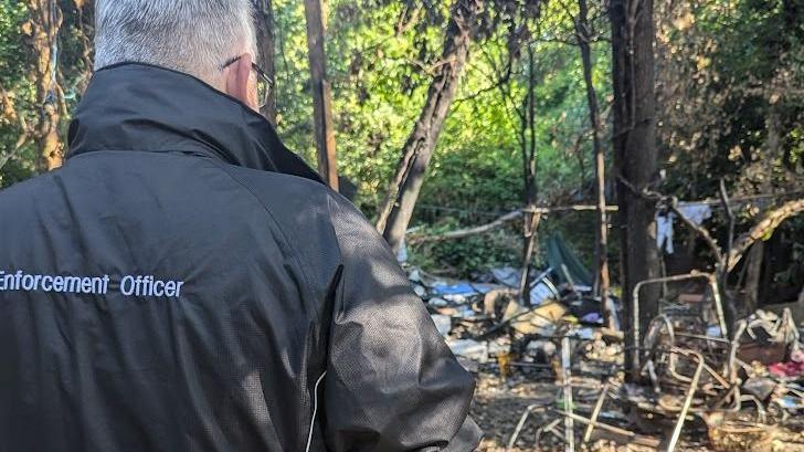 A man wearing a black coat saying 'enforcement officer' on it stands with his back to the camera, looking at piles of rubbish in a wooded area