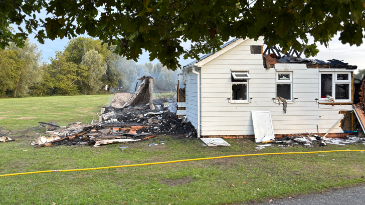 A white building is on the left and stood on a field. The left side of the building has collapsed after a fire which has left a pile of wood. A yellow hose pipe is running across the field.
