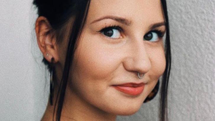 Close up portrait photo of Aimee Walton. She is in her early twenties and has her dark hair tied back with longer strands framing her face at the front. She is smiling and looking to camera.