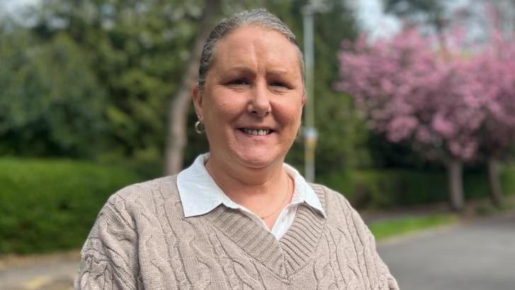 Clare Pretty smiling and looking into the camera. She has grey hair, tied up, and a hoop earring in her left ear. She is wearing a brown knitted v-neck jumper, with a white collared shirt underneath. The background behind her is blurred, but greenery can be seen. On the left, there is a lamp post and a pink blossom tree.