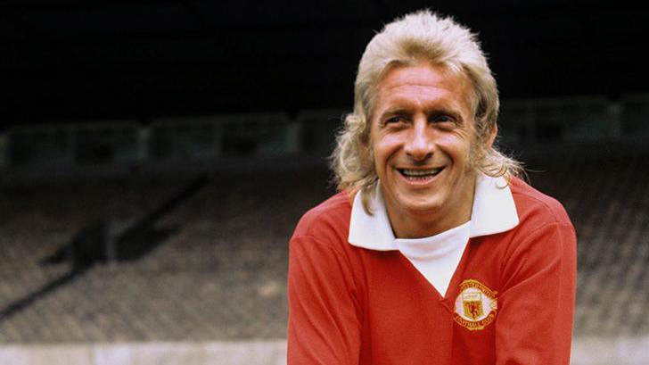 Denis Law, with a blonde mullet, in his early 1970s Manchester United red shirt with extended white V-necked collar