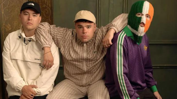 Three men are posing for a photo. Two are wearing caps and one is wearing a green, white and orange balaclava.