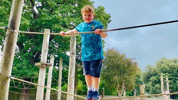Henry standing on a rope in a playground wearing blue shorts and tshirt and blue trainers