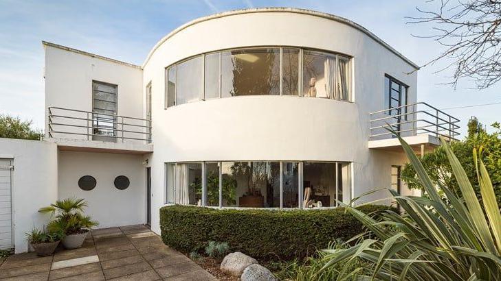 A white flat-roofed two-storey house with a distinctive curved front and windows on each level with a curved hedge in front and paving to a side door. Two steel railing balconies flank the curved facade.