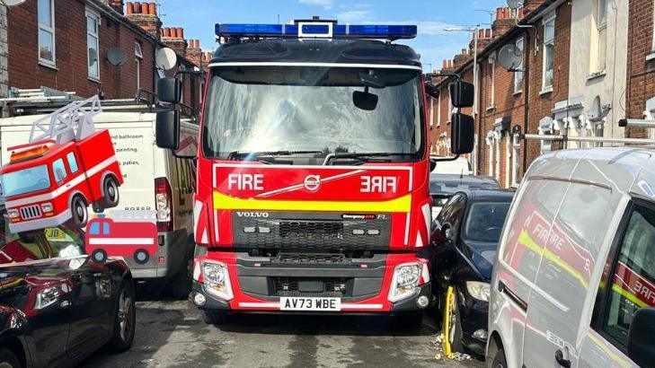 A bright red fire engine attempts to get down a narrow terraced road. It has blue lights on the top and a high-visibility yellow stripe across the front. It is struggling to get past parked vans and a black car which has a yellow clamp on its front right tyre.  