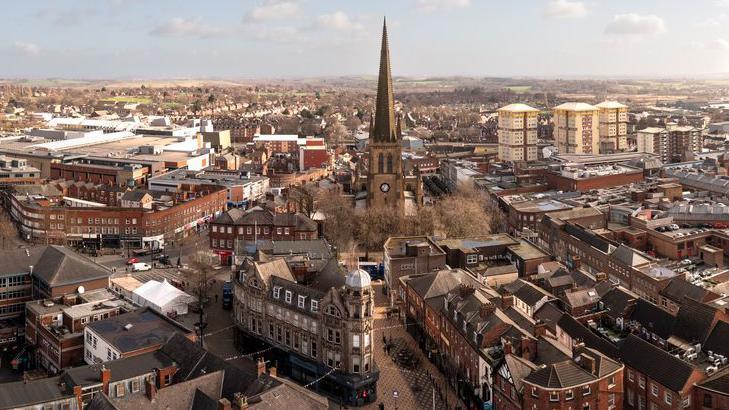 A wide aerial view of Wakefield's city centre surrounded by a dense residential and commercial area. At the centre of the scene stands a tall, pointed church spire made of dark stone, rising prominently above the surrounding buildings. The church beneath the spire is constructed from similar stone and includes a clock face that is visible on the tower.