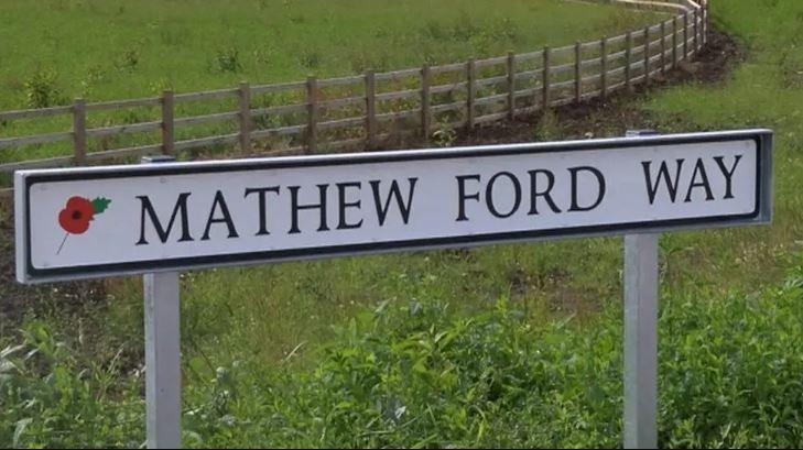 A street sign with a poppy on the left, reading Mathew Ford Way. Grass and a wooden fence can be seen in the background.