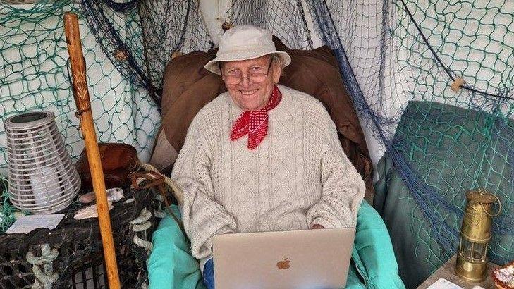 Peter Butler smiling at the camera while sitting in an armchair in a converted shed at the bottom of his garden. He is wearing a white bucket hat, glasses resting on the tip of his nose, a cream knitted jumper, blue jeans and a red handkerchief tied around his neck. There is a laptop resting on his knees on which he writes his stories. He is surrounded by aquatic themed decorations, such as lobster pots, ropes, nets, shells and a gold ship lantern.