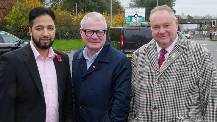 Three men stand next to each other. The man on the left has dark hair and a beard and is wearing a pink shirt and black suit jacket with a poppy. The man in the middle has grey hair and black-rimmed glasses, and is wearing a blue shirt and navy blue coat with a poppy pin. The man on the right has grey hair and is wearing a pink shirt, red tie and checked coat. He has a poppy pin on his lapel. They are all smiling at the camera. Visible behind them is a road sign with a roundabout on, as well as some cars, traffic lights and a road.