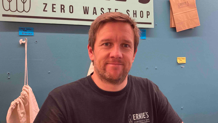 Graham Rutherford in a black t-shirt standing in front of a zero waste shop sign