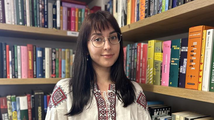 Mariia Kondratiuk is wearing a white dress and is standing in a corner of the library, amongst books in Ukrainian.