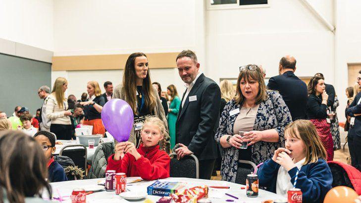 Adults at the IntoUniversity event help chat to pupils as they take part in activities. The children, including two young girls, sit at tables featuring dictionaries, drinks, and balloons.