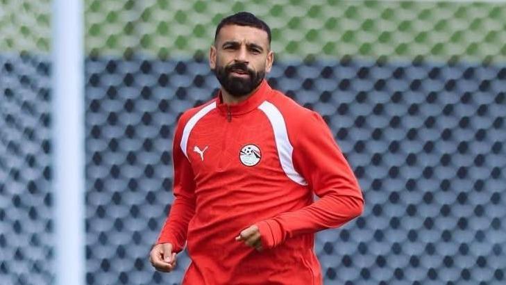 Mohamed Salah, visible from waist up, in training with the Egyptian national team at the World Cup in the USA. Salah wears a red training top with white lines running from armpit to neck on both sides, a white sponsors logo and the crest of the Egyptian FA. The entirety of the background is taken up by a white net with a white goalpost visible to the left of Salah