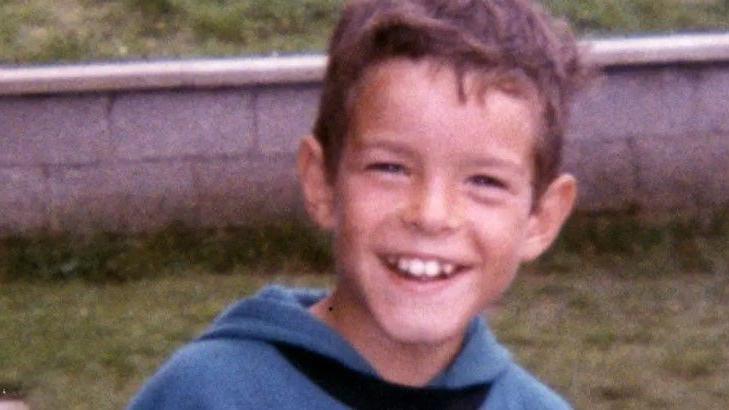 Young boy smiling at the camera. In the background some grass and a concrete wall