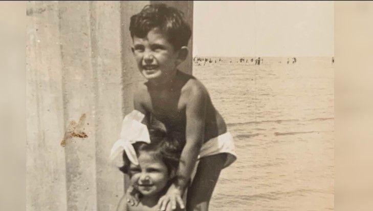 George and his younger sister, pictured as very small children on holiday. They are by the sea and appear to be standing next to a large column, perhaps part of a pier. He is wearing small white swimming trunks, and his sister has a large white both in her hair. Both are smiling and looking away from the camera. 