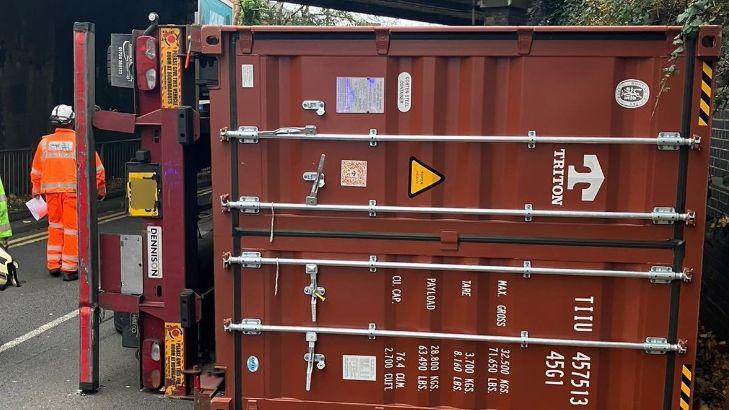 A brown lorry lies on its side in the road and under the bridge. Officials in orange jackets are stood nearby.
