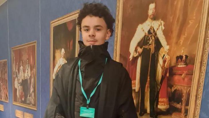 A young man with curly black hair smiling at the camera. He is wearing a black coat and top with a turquoise lanyard on. Behind him are old fashioned portraits of Kings and Queens. 