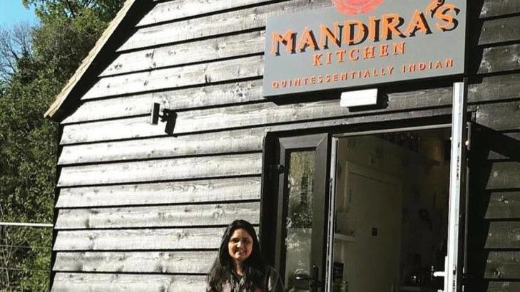 Mandira Sarkar, a South Asian woman with long dark hair, stands outside her restaurant, which is a grey wood panelled building set in countryside.