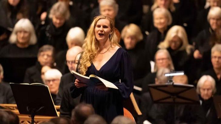 Classical singer Lucy Cox is singing in a choir dressed in black. She is standing up whilst other singers are sat down in the background