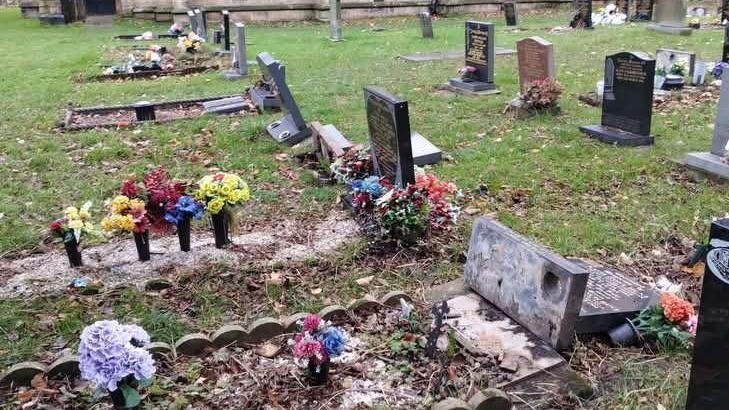 A churchyard with several gravestones and memorials arranged on a grassy area. The ground is scattered with fallen leaves. Some headstones are upright, while others appear tilted or partially fallen. Many graves are decorated with colorful artificial flowers in vases. In the background, a large stone church building with tall arched windows and a dark wooden door is visible.