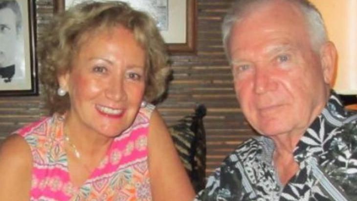 Desmond and Mary Byrne smiling into the camera. Mary has blonde, short hair and is wearing a floral pink top and is wearing pink lipstick. Desmond has grey hair and is wearing a green, patterned shirt.