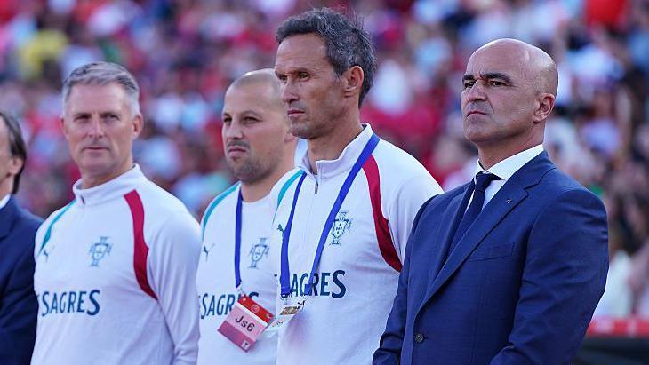 Richie Evans in a white portugal training top stands in an anthem line up of four men, with manager Roberto Martinez on the right nearest camera wearing a suit