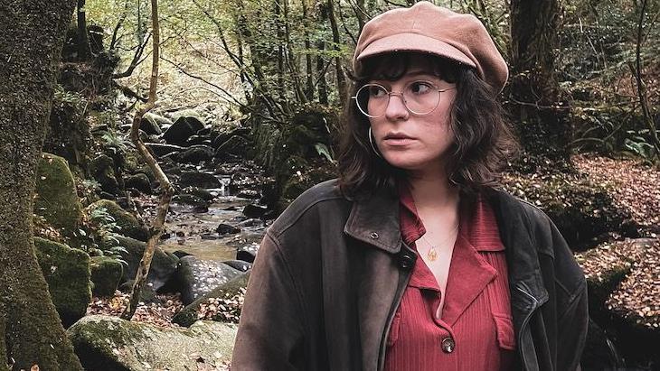 Alice, a woman with shoulder-length brown hair, is wearing a beige cap and large glasses. She looks to one side and is standing in a glen, with a shallow stream behind her.