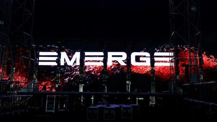 The graphics screen on the stage at the Emerge event spells out the word 'Emerge' in white block letters with a red liquid effect behind it. 