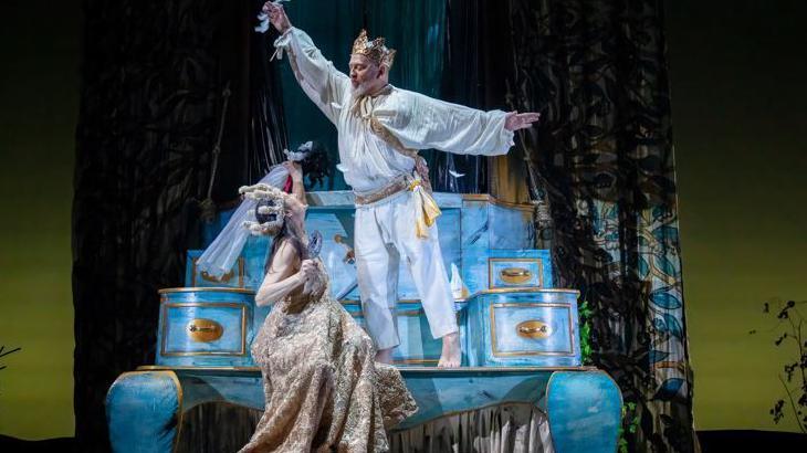 Geoff Hopson is in a white and gold costume looking down on the queen. He is wearing a gold crown and his arms are outstretched. The stage set is a huge representation of an ornate, blue and gold Regency style chest of drawers.