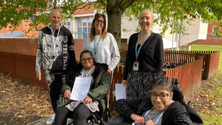 Three students, two in wheelchairs, and two members of staff in front of a garden area which is surrounded by a waist-high fence