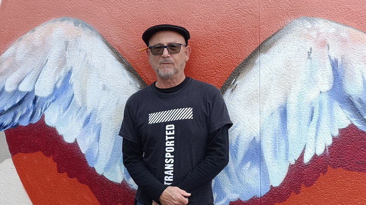 Nick Jones is wearing a black t-shirt and is standing in front of a wall with painted white angel wings on it.