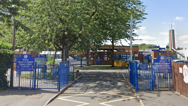 The front gates of Bilston primary school. There is a large tree on the left, and the school is in the background. To the right, inside the gates, is a car park