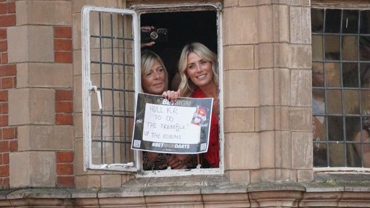 Two women hanging out of a window. One of them has a sign that reads Hull KR to do the treble. Up the Robins.