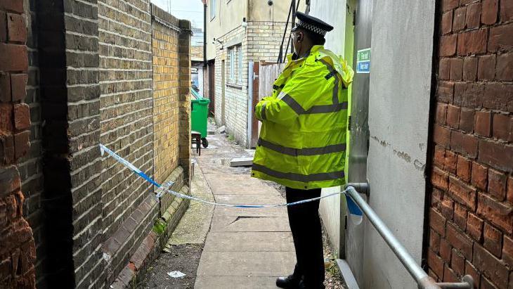 A police officer wearing a black hat and hi-vis jacket. He is standing behind blue and white police tape down an alleyway, which has brick walls either side of it and a wheelie bin in the background.