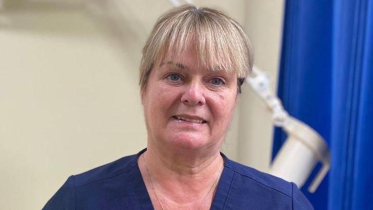 Dr Frances O'Hagan has blonde hair with a full fringe, with her hair tied back. She is smiling at the camera and is wearing navy scrubs. Behind her is an examination bed and a blue privacy curtain but they are both blurred slightly.