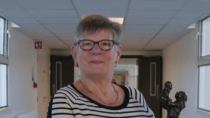 Woman with glasses and cream and black-stripped top holds her award - a sculpture of a midwife holding a baby in bronze and wood. She is stood in a hospital corridor