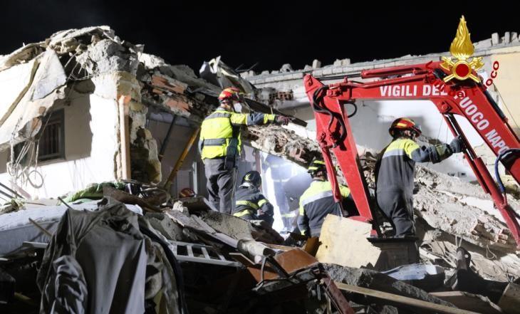 Rescue workers stand on top of rubble, working alongside an excavator