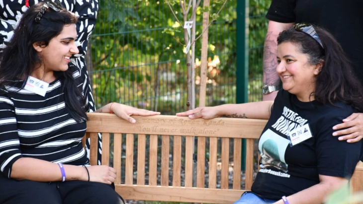 Two women sit on a bench in a garden and smile. The bench has an inscription on it, which says: "In memory of Ronan Kanda. You'll never walk alone."