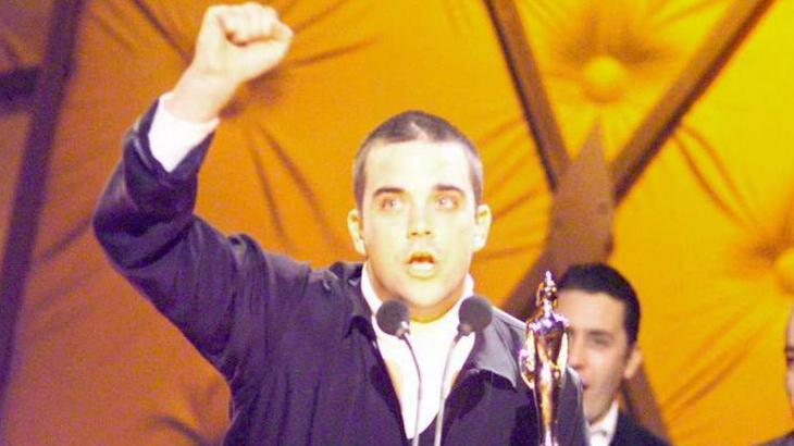 A younger Robbie Williams, with a buzz cut, stands up on stage with his fist in the area as he receives a Brit Award for his single Angels.