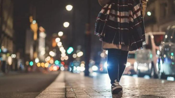 An anonymous woman walks down a street at night, with black and white sneakers, with black tights, and a beige coat.  To her left is a row of vehicles and to the right a row of buildings - both are blurred out.