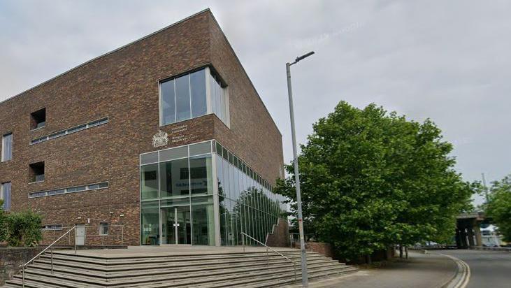 Photograph of the outside of Newport Magistrates Court. Stairs can be seen leading up to the entrance on the corner of the building. Large windows cover two floors on the right hand side. A road can be seen on the right, along with some large trees in Newport, south Wales.