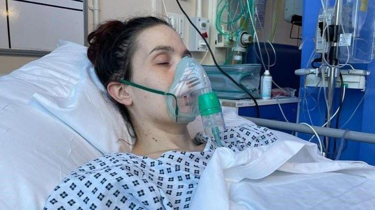 Natalie is lying in a hospital bed wearing a hospital gown and receiving oxygen through a face mask. Medical equipment, monitors and tubing are visible around the bed, indicating a high-dependency or intensive care setting. The patient is covered with white bedding, and a scar can be seen on one arm