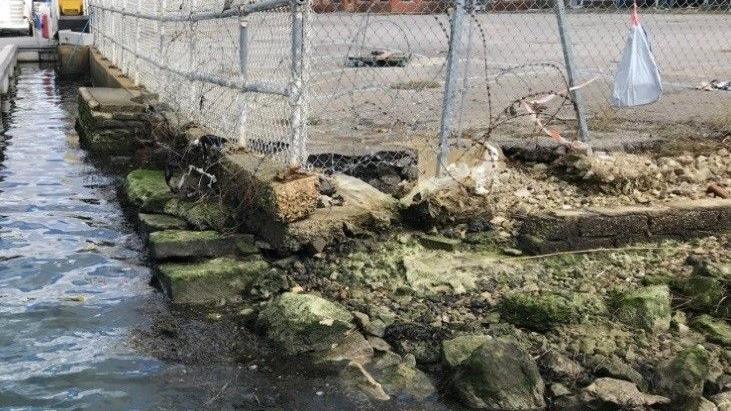 Crumbling sea walls surrounding a fenced tarmac area. The water is almost up to the same level as the ground.